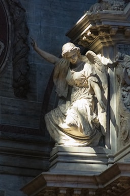 A stone statue of an angel is gracefully positioned with wings partially spread and one arm raised. The figure is adorned in flowing robes, highlighted by soft light creating a dramatic contrast against the shadowed background. Architectural details surround the statue, enhancing its majestic presence.