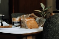 A loaf of bread partially wrapped in plastic sits on a wooden table next to an open jar of peanut butter, a plate with a slice of bread, and a knife. A moka pot is visible on the left and there is a plant in the background.