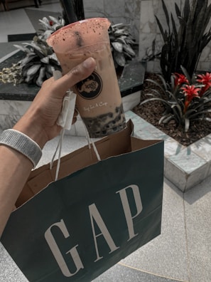 A hand is holding a cup of bubble tea with a lid and a straw, adorned with a branded logo. The same hand is carrying a shopping bag with the GAP logo. The setting appears to be indoors, with decorative plants and tiled flooring visible in the background.