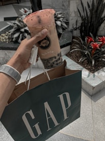 A hand is holding a cup of bubble tea with a lid and a straw, adorned with a branded logo. The same hand is carrying a shopping bag with the GAP logo. The setting appears to be indoors, with decorative plants and tiled flooring visible in the background.