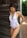 A woman wearing a white one-piece swimsuit with lace-up details on the sides stands next to a rustic wooden wall, with greenery visible in the background. She is looking down and wearing drop earrings.