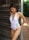 A woman wearing a white one-piece swimsuit with lace-up details on the sides stands next to a rustic wooden wall, with greenery visible in the background. She is looking down and wearing drop earrings.