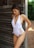 A woman wearing a white one-piece swimsuit with lace-up details on the sides stands next to a rustic wooden wall, with greenery visible in the background. She is looking down and wearing drop earrings.