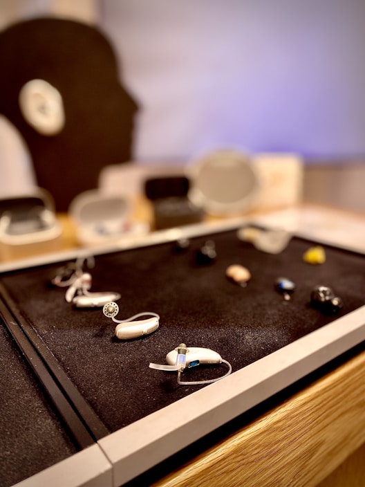 Several hearing aids are arranged on a black display surface. In the background, a silhouette of a human head is visible, suggesting a focus on auditory devices.