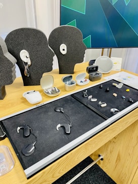 A wooden table displays various hearing aid devices in their respective cases. Several head-shaped models showcase the devices. In the background, a screen is visible with abstract green and blue graphics.