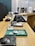 A row of various small electronic devices and objects is laid out on a wooden surface. The background is slightly blurred, highlighting the focus on the items. Some items are in clear plastic containers, and there are labels visible on certain objects.
