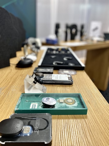 A row of various small electronic devices and objects is laid out on a wooden surface. The background is slightly blurred, highlighting the focus on the items. Some items are in clear plastic containers, and there are labels visible on certain objects.