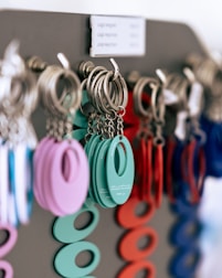 A close-up of multiple keychains hanging on a display rack. The keychains are circular with an oval cutout in the center, coming in various colors including teal, purple, red, and blue. The keychains are attached to metal rings, and they appear to be organized by color.
