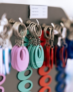 A close-up of multiple keychains hanging on a display rack. The keychains are circular with an oval cutout in the center, coming in various colors including teal, purple, red, and blue. The keychains are attached to metal rings, and they appear to be organized by color.