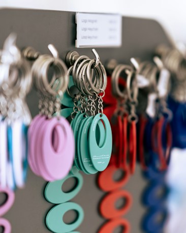 A close-up of multiple keychains hanging on a display rack. The keychains are circular with an oval cutout in the center, coming in various colors including teal, purple, red, and blue. The keychains are attached to metal rings, and they appear to be organized by color.