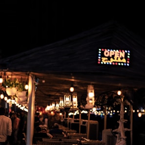 A dimly lit outdoor dining area features hanging lanterns and string lights creating a warm, inviting ambiance. A wooden structure with hanging plants adds a cozy touch. An 'OPEN' sign with colorful lights is prominently displayed. Patrons are seen seated and walking around, suggesting a lively, social atmosphere.