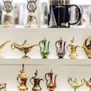 Various intricately designed miniature Arabian teapots and magic lamps are displayed on a white shelf. These ornate items feature colorful patterns in gold, green, and purple. In the background, a black mug with an image of a skyscraper and 'Dubai' written on it is also visible.