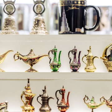 Various intricately designed miniature Arabian teapots and magic lamps are displayed on a white shelf. These ornate items feature colorful patterns in gold, green, and purple. In the background, a black mug with an image of a skyscraper and 'Dubai' written on it is also visible.