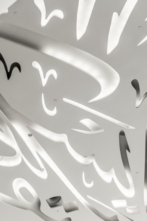 An abstract ceiling design featuring intricate, sculptural cutouts with soft, curved shapes. Gentle lighting emanates from within the cutouts, creating a contrast between illuminated areas and shadows. The overall appearance is modern and artistic, with a sleek and clean design.