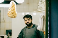 A man stands in what appears to be a kitchen or bakery setting. He has a serious expression on his face and is wearing a dark, simple top. To the left, there is a large piece of hanging bread. The background shows tiled walls and some kitchen equipment.
