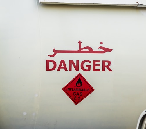 Large bold red text written in both Arabic and English against a light background, indicating a warning with the word 'DANGER'. Below is a red diamond-shaped sign with a symbol of a flame and the words 'INFLAMMABLE GAS 2.1'.