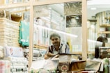 A person stands inside a fabric or textile store, surrounded by neatly stacked fabric rolls and packages. The shop is well-lit, with various fabrics displayed on shelves. The individual is looking towards the camera with a relaxed yet observant expression.