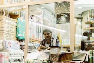 A person stands inside a fabric or textile store, surrounded by neatly stacked fabric rolls and packages. The shop is well-lit, with various fabrics displayed on shelves. The individual is looking towards the camera with a relaxed yet observant expression.