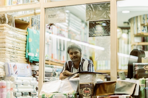 A person stands inside a fabric or textile store, surrounded by neatly stacked fabric rolls and packages. The shop is well-lit, with various fabrics displayed on shelves. The individual is looking towards the camera with a relaxed yet observant expression.