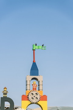 A colorful LEGO structure featuring a tower with a blue pointed top, a yellow and orange base, and large gray gears at the bottom. Two LEGO minifigures are present: one dressed as a firefighter on the left and another in a red suit standing within the tower arches. A green flag flies at the top, with two birds perched on it. The background is a clear blue sky.
