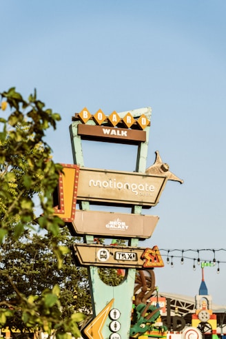 A tall signpost with multiple rectangular signs pointing in different directions, indicating locations such as 'Boardwalk,' 'Motiongate Dubai,' and 'Neon Galaxy Indoor Entertainment.' The signpost is surrounded by foliage, with a clear blue sky in the background.