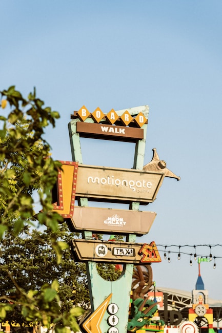 A tall signpost with multiple rectangular signs pointing in different directions, indicating locations such as 'Boardwalk,' 'Motiongate Dubai,' and 'Neon Galaxy Indoor Entertainment.' The signpost is surrounded by foliage, with a clear blue sky in the background.