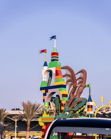 Colorful LEGO-style structure resembling a tall castle with two small flags at the top. A large, detailed green dragon is wrapped around part of the structure. Palm trees and the roof of a nearby building are visible in the background, and a portion of a vehicle can be seen in the foreground.