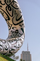 A futuristic building with large, flowing Arabic calligraphy etched onto its metallic exterior, set against a clear blue sky. In the background, a tall, pointed skyscraper stands prominently.