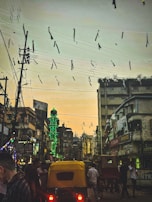 A bustling street scene at dusk with a glowing green minaret contrasting against the evening sky. Overhead, tangled electric wires are festooned with streamers, while below, auto rickshaws navigate through a crowd of people amidst tall urban buildings adorned with colorful signs.