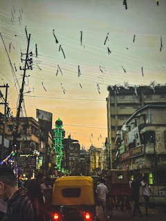 A bustling street scene at dusk with a glowing green minaret contrasting against the evening sky. Overhead, tangled electric wires are festooned with streamers, while below, auto rickshaws navigate through a crowd of people amidst tall urban buildings adorned with colorful signs.