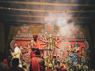 A traditional religious setup featuring intricately decorated statues of deities, with figures in ceremonial attire performing a ritual. Smoke rises amidst the vibrant decorations, adding a mystical atmosphere to the scene.