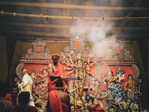 A traditional religious setup featuring intricately decorated statues of deities, with figures in ceremonial attire performing a ritual. Smoke rises amidst the vibrant decorations, adding a mystical atmosphere to the scene.