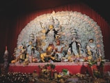 A detailed and ornate display features a group of Hindu deities, prominently showcasing the goddess Durga in the center with multiple arms, each holding a different object. She is flanked by other gods and goddesses, each intricately decorated with vibrant garments and jewelry. The backdrop is an elaborate design of intricate patterns and motifs. Offerings and ritual objects are placed at the forefront of the display.
