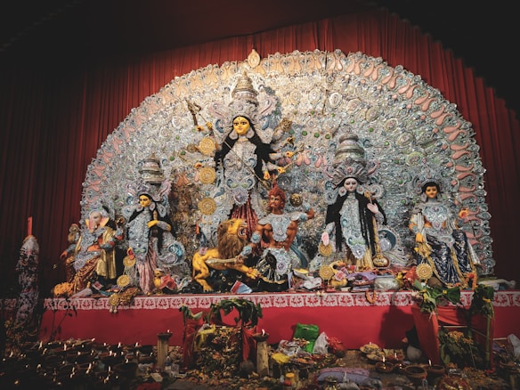 A detailed and ornate display features a group of Hindu deities, prominently showcasing the goddess Durga in the center with multiple arms, each holding a different object. She is flanked by other gods and goddesses, each intricately decorated with vibrant garments and jewelry. The backdrop is an elaborate design of intricate patterns and motifs. Offerings and ritual objects are placed at the forefront of the display.