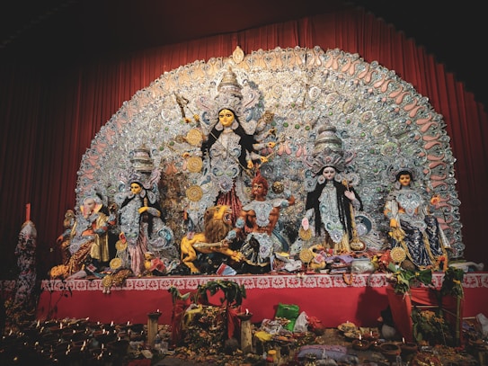A detailed and ornate display features a group of Hindu deities, prominently showcasing the goddess Durga in the center with multiple arms, each holding a different object. She is flanked by other gods and goddesses, each intricately decorated with vibrant garments and jewelry. The backdrop is an elaborate design of intricate patterns and motifs. Offerings and ritual objects are placed at the forefront of the display.