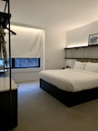 A modern hotel room features a neatly made double bed with white linens and a stylish headboard. There is a framed picture placed on a shelf above the bed, and a window with a closed blind, allowing a bit of natural light to filter in. The room is minimally decorated, with a neutral color palette and a clean, uncluttered feel. A small hanging rack with some clothing items is visible on the left.