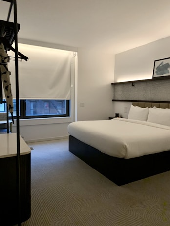 A modern hotel room features a neatly made double bed with white linens and a stylish headboard. There is a framed picture placed on a shelf above the bed, and a window with a closed blind, allowing a bit of natural light to filter in. The room is minimally decorated, with a neutral color palette and a clean, uncluttered feel. A small hanging rack with some clothing items is visible on the left.