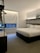 A modern hotel room features a neatly made double bed with white linens and a stylish headboard. There is a framed picture placed on a shelf above the bed, and a window with a closed blind, allowing a bit of natural light to filter in. The room is minimally decorated, with a neutral color palette and a clean, uncluttered feel. A small hanging rack with some clothing items is visible on the left.