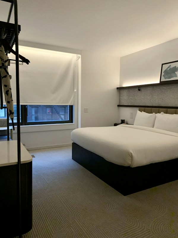 A modern hotel room features a neatly made double bed with white linens and a stylish headboard. There is a framed picture placed on a shelf above the bed, and a window with a closed blind, allowing a bit of natural light to filter in. The room is minimally decorated, with a neutral color palette and a clean, uncluttered feel. A small hanging rack with some clothing items is visible on the left.