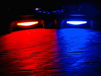 Two video game controllers are placed side by side on a wooden surface in a dark environment, emitting vibrant lights from their light bars&mdash;one glowing red and the other blue. The colors reflect off the surface, creating a striking contrast between the red and blue hues.