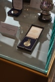 A display case with several medals, one prominently featured in an open black case lined with blue fabric. The medal is gold, featuring intricate engravings. Nearby, a plaque notes 'Micky Hazard's F.A. Cup winners medal 1981/1982'. The setup is elegant, with reflective surfaces highlighting the polished silver of a nearby trophy.