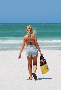 A person with long hair, wearing a black bikini top and denim shorts, walks away on a sandy beach toward the ocean. They hold a colorful tote bag in one hand and are barefoot. The sea is calm with gentle waves, and the sky above is clear with a deep blue hue.