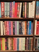 A collection of books neatly arranged on a wooden bookshelf. The books are of various colors and sizes, with some titles clearly visible, such as 'Devil Dogs', 'The Woman in the Window', and 'Circe'. The overall atmosphere suggests a well-organized personal or public library section, showcasing a diverse range of genres.