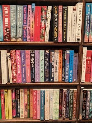 A collection of books neatly arranged on a wooden bookshelf. The books are of various colors and sizes, with some titles clearly visible, such as 'Devil Dogs', 'The Woman in the Window', and 'Circe'. The overall atmosphere suggests a well-organized personal or public library section, showcasing a diverse range of genres.