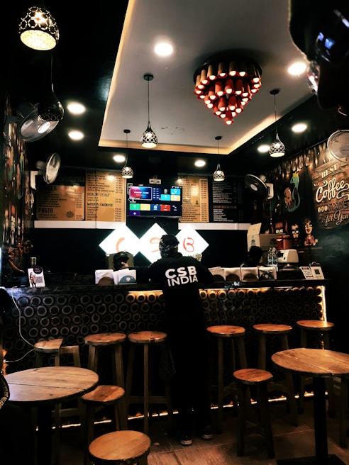 A cozy coffee shop interior with warm lighting and wooden furniture. The counter features circular decorative elements, and a person wearing dark clothing stands at the counter. Overhead, several decorative lights hang from the ceiling, and a digital menu is displayed behind the counter.