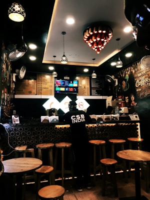 A cozy coffee shop interior with warm lighting and wooden furniture. The counter features circular decorative elements, and a person wearing dark clothing stands at the counter. Overhead, several decorative lights hang from the ceiling, and a digital menu is displayed behind the counter.