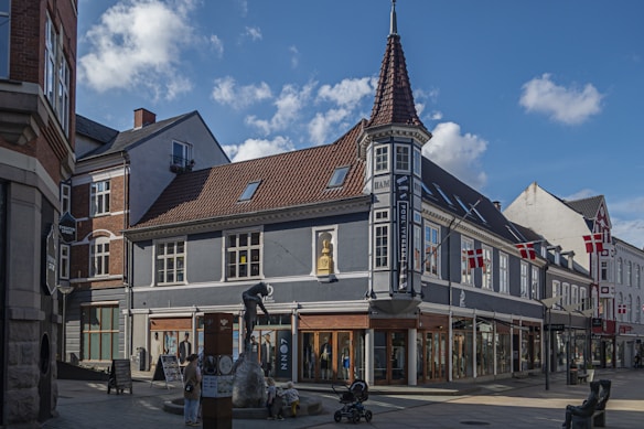 A quaint street corner in a European town, featuring a building with a distinctive turret and multiple Danish flags. The building has a muted color palette with a gray fa&ccedil;ade and a brown tiled roof. Commercial signs are visible, indicating retail spaces on the ground floor. There are people casually walking and interacting, and a sculpture of a man climbing a rock is prominently displayed in front of the building.