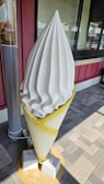 A large ice cream sculpture displayed outside a store. The sculpture features a swirled soft serve ice cream with a smooth texture, placed in a cone. The cone is secured to the ground by concrete blocks and rope. It is positioned on a tiled pavement in front of a building with red and gray tones.