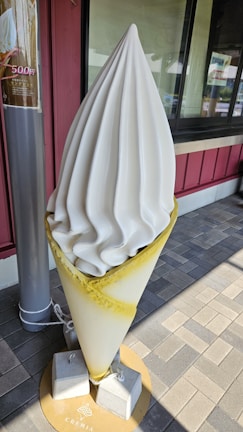 A large ice cream sculpture displayed outside a store. The sculpture features a swirled soft serve ice cream with a smooth texture, placed in a cone. The cone is secured to the ground by concrete blocks and rope. It is positioned on a tiled pavement in front of a building with red and gray tones.