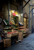 A quaint street market stall displaying a variety of fresh fruits and vegetables. There are wooden crates filled with colorful produce such as tomatoes, peppers, bananas, and lettuce. The cobblestone street is wet, possibly from rain, adding a fresh atmosphere.